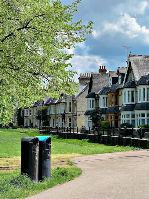 A row of Victorian-style terraced houses with bay windows and slate roofs, situated behind a black metal fence. In the foreground, there are two black public litter bins positioned on a grassy verge beside a paved footpath. A large, leafy tree with bright green foliage extends its branches over the scene from the left side, partially shading the area. The background shows a partly cloudy sky with patches of blue, adding natural light to the residential setting. The image reflects a typical urban or suburban street scene where rubbish collection or waste disposal might be managed through community bins or external collection services, with the presence of public litter bins complementing private waste handling options occasionally used in such neighbourhoods. The scene’s detail emphasizes a clean, well-maintained environment, consistent with waste management involving both community and private collection practices.