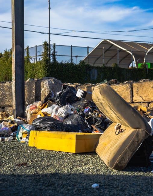 A pile of discarded rubbish consisting of black plastic bin bags, a yellow plastic tray, a large worn beige car tire, and various small debris, all resting on a gravel surface near a stone retaining wall. The rubbish appears to be gathered for collection outside a residential or commercial area, with a background featuring a chain-link fence, greenery, and a partially visible covered structure. The scene is illuminated by natural daylight, with shadows cast by the objects, indicating clear weather. This visual context illustrates an instance of waste accumulation suitable for private rubbish removal services such as those offered by Rubbish Collection Roehampton, especially involving on-site clearance of mixed waste materials that require alternative disposal methods outside regular council collection.